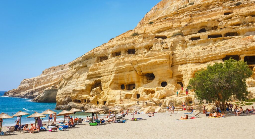 Best Sandy Beaches in Crete - 7. Matala Beach Photo. 

## **Matala Beach Location**

**Matala Beach** is located on the southern coast of **Crete**, in the **Heraklion** region. Situated about **70 kilometers south of Heraklion** city, Matala is a charming beach destination known for its beautiful scenery, crystal-clear waters, and historical significance.

The beach is located near the village of **Matala**, which is famous for its natural caves carved into the surrounding cliffs. These caves were once used as ancient tombs and later became popular with the counterculture movement in the 1960s. Today, Matala Beach is a popular destination for both history enthusiasts and beach lovers.

### **How to Get to Matala Beach**

1. **From Heraklion**: To reach **Matala Beach** from Heraklion, take the **E70 road** heading south. After about an hour and 15 minutes (roughly 70 km), you will reach the village of Matala. Follow the signs for the beach, and you'll find a large parking area nearby.

2. **From the Village of Matala**: The beach is a short walk from the village center. Once you reach the village, the path to the beach is easy to find, and it’s only a few minutes’ walk to reach the shore.

3. **Access and Facilities**: Matala Beach is easily accessible by car, and parking is available near the beach. It is also well-equipped with facilities like cafes, restaurants, and sunbeds, making it a convenient destination for tourists.

Matala Beach is easily accessible and remains a beautiful destination with a unique blend of history, natural beauty, and modern-day comforts.