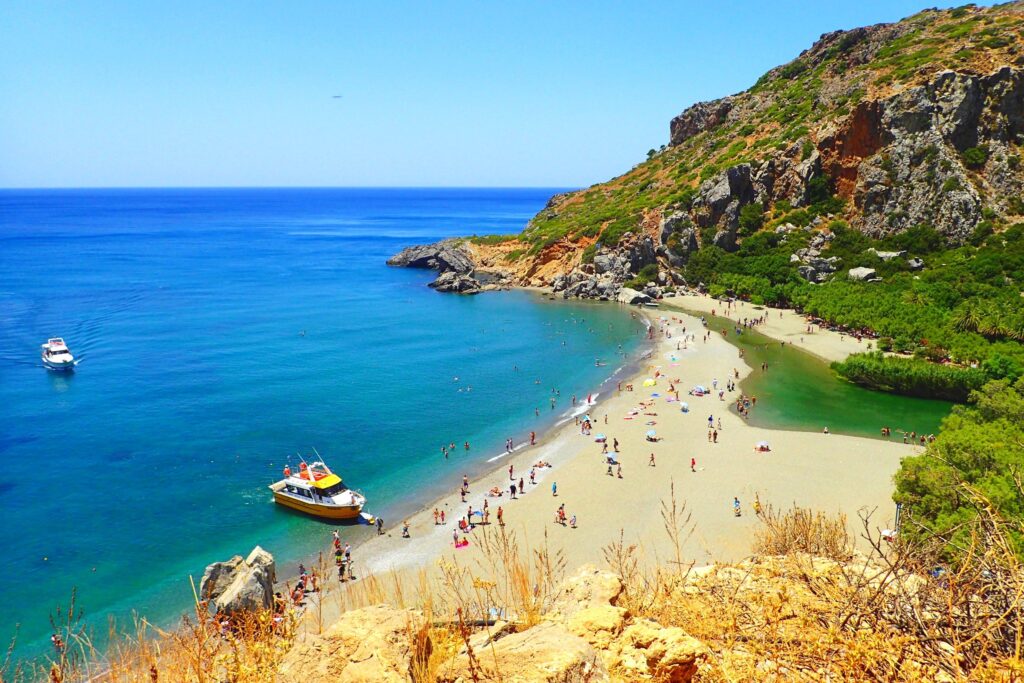 Best Sandy Beaches in Crete - 8. Preveli Beach. ## **Preveli Beach: A Hidden Paradise in Southern Crete**

**Preveli Beach**, located on the southern coast of **Crete**, is one of the island’s most iconic and beautiful beaches. Known for its lush palm forest, crystal-clear waters, and serene environment, Preveli offers a unique blend of natural beauty, history, and tranquility. It is one of the most sought-after destinations for those looking to experience both a relaxing beach day and an adventure in nature.

### **Why Preveli Beach is Famous**

**Preveli Beach** stands out for its stunning natural landscape. The beach is set in a picturesque location, where the **Kourtaliotiko River** meets the sea, forming a small lagoon and creating an idyllic spot for swimming and relaxation. The most unique feature of the beach is the **palm forest**, **one of the few in Europe**, which grows right at the edge of the beach, offering shade and a tropical atmosphere.

This combination of river, palm trees, and the Libyan Sea creates a magical setting that feels far removed from the more crowded tourist beaches on the northern coast of Crete. Preveli has not only become a favorite for beach lovers but also for those interested in hiking, photography, and exploring Crete's diverse landscapes.

### **The Best Activities at Preveli Beach**

1. **Swimming and Sunbathing**: Preveli Beach is perfect for those looking to enjoy a peaceful swim in beautiful surroundings. The shallow lagoon formed by the river’s mouth is calm and warm, making it great for swimming, especially for families with children. The beach's clear blue waters are ideal for relaxing swims or simply enjoying the beach.

2. **Exploring the Palm Forest**: The palm forest surrounding Preveli Beach is one of the most remarkable features of the area. The **Cretan date palms** offer much-needed shade during hot summer days, and the sight of the palms next to the beach creates a tropical, tranquil vibe. You can walk through the forest and enjoy the unique ecosystem that surrounds you.

3. **Hiking**: For hiking enthusiasts, there are several hiking routes leading to and from Preveli Beach. One of the most popular trails is the path that descends from **Preveli Monastery** down to the beach. The route offers stunning panoramic views of the sea, the surrounding cliffs, and the river. The hike takes about **30 to 40 minutes** and is moderately challenging.

4. **Photography**: With its dramatic landscapes, crystal-clear waters, and unique combination of palm trees and mountains, Preveli Beach is a photographer's paradise. Whether you are capturing the beauty of the river meeting the sea, the rich green palm trees, or the view from the cliffs above, Preveli offers plenty of opportunities for stunning photography.

5. **Kayaking and Canoeing**: The calm waters of the lagoon are perfect for kayaking or canoeing. Renting a kayak is a great way to explore the coastline from the water, as you can paddle along the river or explore the surrounding areas and hidden coves.

### **How to Get to Preveli Beach**

1. **From Rethymno**: Preveli Beach is located about **35 kilometers south of Rethymno**, and it is easily accessible by car. The drive takes around **45 minutes to 1 hour**. From Rethymno, take the **E70** road south, following the signs to **Plakias**. From Plakias, continue along the road that leads to Preveli Beach, where you will find a parking area.

2. **From the Parking Area**: There are two main access points to the beach. The easiest way is to take the **road to the parking lot**, which is located just above the beach. From there, you can either walk down a fairly steep path (about 10-15 minutes) or take a **hiking route** from the nearby **Preveli Monastery**, which takes about 30-40 minutes.

3. **Public Transport**: Public buses run to **Plakias** from **Rethymno**, but from there, you would need to rent a car or take a taxi to reach the beach.

### **Best Time to Visit Preveli Beach**

The best time to visit Preveli Beach is during the **spring (April to June)** and **early autumn (September to October)**. During these months, the weather is warm, but the crowds are smaller, and the area remains peaceful. The summer months (July and August) can be busier, especially with tourists heading to the beach, so visiting early in the morning or later in the afternoon is recommended for a more serene experience.

### **Conclusion**

**Preveli Beach** is a breathtaking destination that combines natural beauty, tranquility, and adventure. Its unique palm forest, pristine waters, and peaceful surroundings make it one of Crete’s most beloved beaches. Whether you’re looking to relax by the sea, hike through nature, or explore its rich ecosystem, Preveli offers a perfect escape from the bustling crowds of the more tourist-heavy areas of Crete. With its stunning views and serene atmosphere, Preveli is truly one of Crete's hidden gems.